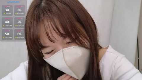 Video of a young Asian woman with long, straight brown hair, wearing a white surgical mask, covering her nose and mouth, in a sterile white room.