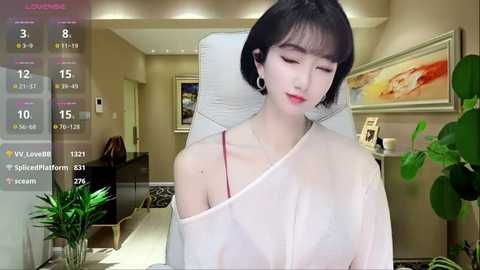 Media: A video of an East Asian woman with short black hair, wearing a sheer white dress, sitting in a modern living room with beige walls and framed art.