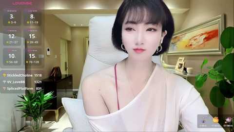 Video of an East Asian woman with a bob haircut, fair skin, and large earrings, wearing a white off-shoulder top, sitting on a white chair in a modern living room.