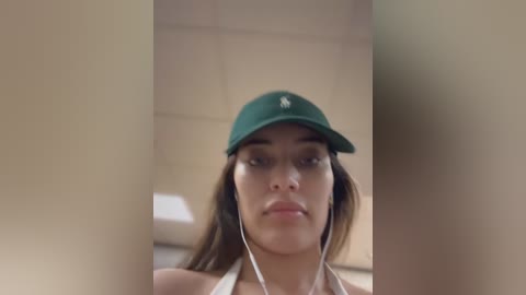 Media: Video of a young woman with light brown skin, wearing a green baseball cap, headphones, and a white halter top, standing in a beige tiled bathroom.