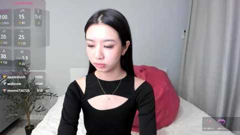 Media: A video of a young East Asian woman with long black hair, wearing a black top with a keyhole cutout, sitting on a bed with a red pillow. The room has a digital overlay showing social media metrics.