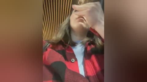 Media: Video of a young woman with light skin, long brown hair, and a red and black plaid shirt, covering her face with her right hand, sitting in a wooden chair with vertical slats.