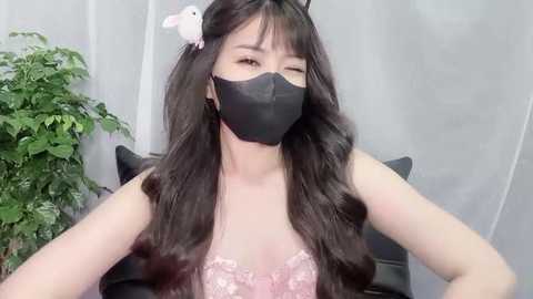 Media: Video of an Asian woman with long, dark hair, wearing a black face mask and a pink lace bra, against a white curtain background with a potted plant.