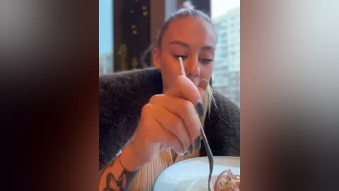 Media: Video of a young woman with light skin, long blonde hair in a ponytail, eating with chopsticks at a restaurant table. She wears a dark jacket and has a tattoo on her left wrist. The background shows a blurred cityscape.