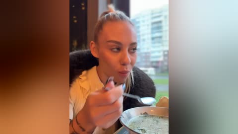 Media: A video of a woman with blonde hair in a ponytail, wearing a black fur-trimmed coat, eating soup at a table. Background shows a blurred cityscape through a window.