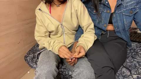 Media: Video of two women sitting on a bed; one in a beige zip-up hoodie and blue jeans, the other in a denim jacket over a black top and dark pants.