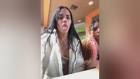 Media: Video of a woman with long black hair, wearing a white jacket, sticking out her tongue, and a woman with curly hair in the background.