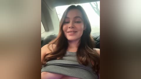 Media: A video shows a young woman with long brown hair, wearing a grey tank top, grinning while seated in a car, with blurry windows and a green tree outside.