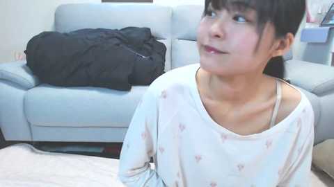 Video of an Asian woman with straight black hair, wearing a light-colored pajama top, sitting indoors, looking up. Background features a light gray couch with a black jacket and a wall-mounted calendar.