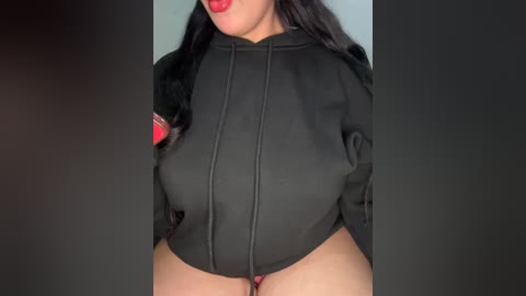 Media: Video of a woman with long black hair, wearing a black zip-up jacket with red accents, revealing her large breasts and exposed pubic area, against a dark background.