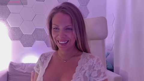 Media: Video of a smiling, light-skinned woman with long blonde hair, wearing a white lace top, seated in a white leather chair. Background features a hexagonal patterned wall and soft lighting.