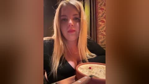 Media: Video of a blonde woman with long hair, wearing a black top, holding a bowl of creamy soup, in a dimly lit, cozy room with patterned wallpaper.