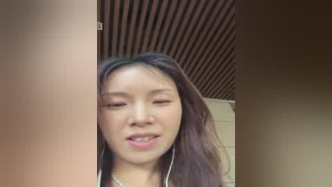 Media: A video of a young Asian woman with long, straight hair, wearing a white necklace and headphones. She has a light skin tone and is smiling. The background features a wooden ceiling and beige wall.