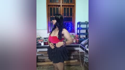 Media: Video of a curvy, light-skinned woman with long black hair, wearing a red top and black ruffled skirt, standing in a cluttered room with wooden cabinets and shelves.