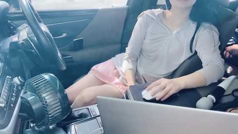 Media: Video of an East Asian woman in a car, wearing a grey shirt and pink skirt, typing on a laptop, with a fan and black bag beside her.