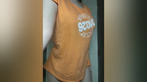 Media: Video of a woman with a fair complexion, wearing an orange sleeveless t-shirt with \"Brooklyn\" text and a round logo, standing indoors with blurred background.