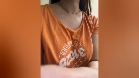 Media: Video of a woman wearing an orange \"Brooklyn\" t-shirt with a circular logo, partially obscured by a blurred, orange background. She has a light skin tone and is indoors.