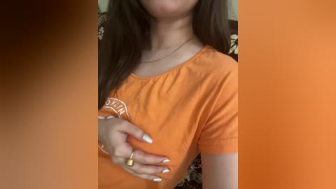 Media: Video of a woman with long dark hair, wearing an orange T-shirt, touching her chest with her hand, against an orange backdrop.