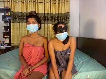 Media: Video of two women sitting on a bed, wearing masks, pink and grey towels, in a modest, dimly-lit bedroom with yellow curtains.