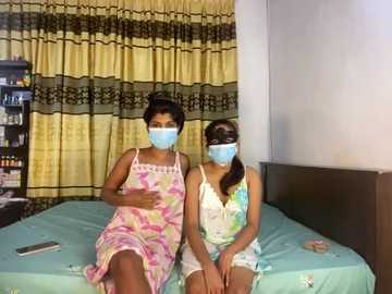 Media: Video of two young women wearing face masks, sitting on a bed with light blue sheets, in a room with yellow curtains and a shelf of toiletries.