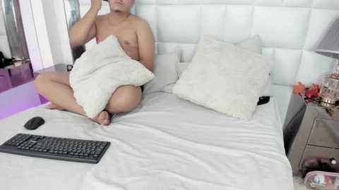 Media: A video of a shirtless Asian man, with a medium build, lying on a white bed with pillows, holding a white pillow, wearing black pants, on a white headboard.