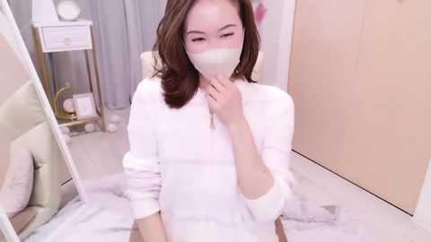 Media: Video of an Asian woman with fair skin, wearing a white mask, white robe, and sitting on a white bed, in a modern, minimalist bedroom with a white dresser and mirror.