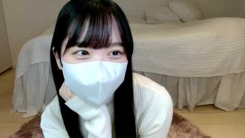 Media: A video of an Asian woman with straight black hair, wearing a white face mask, light sweater, and black suspenders, kneeling on a beige rug in a minimalist bedroom with a white bed.