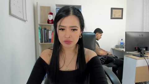 Media: Video of a young woman with long black hair, wearing a black off-shoulder top, seated in an office. A man sits behind a desk in the background.