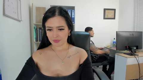 Media: Video of a Latina woman with long black hair and tattoos, wearing a black off-shoulder top, sitting in an office. A man works in the background.