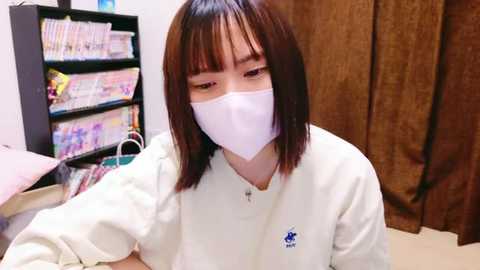 Media: Video of an Asian woman with shoulder-length brown hair, wearing a white mask and white polo shirt, sitting in a room with a bookshelf and brown curtains.