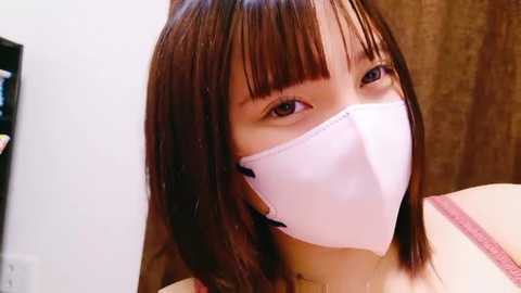 Media: A video of an East Asian woman with straight brown hair and bangs, wearing a white surgical mask and a pink bra. She has a neutral expression and is indoors with a wooden door and part of a black bookshelf visible in the background.