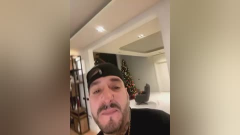 Media: Video of a man with a black baseball cap, facial hair, and a tattoo on his neck, standing in a modern, dimly lit living room with a Christmas tree and white furniture.