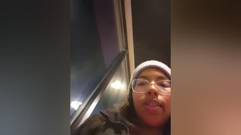 Media: Video of a young woman with light brown skin, wearing a white beanie, glasses, and a camouflage jacket, smiling as she peers through a dark window.