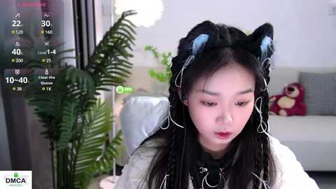 Media: Video of an East Asian woman with long black hair, wearing cat ears, sitting indoors. She has a pale complexion and is wearing a white sweater. The background features a potted fern, a red cushion, and a white couch.