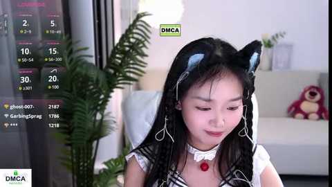 Media: Video of a young East Asian woman in a cat costume with black ears, wearing a white blouse, smiling, in a modern living room with a plant, plush toys, and a digital clock displaying 10:15.