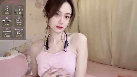 Media: Video of a young Asian woman with fair skin, wearing a pink tank top, sitting on a bed with beige sheets. She has long brown hair in a loose bun, and her expression is neutral.