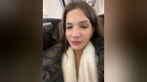 Media: Video of a young woman with light brown skin, long brown hair, wearing a black leather jacket and white scarf, seated in a car with blurred surroundings.