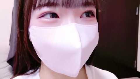 Media: Video of an East Asian woman with straight, dark brown hair and light skin, wearing a white face mask and a white top. Her large, almond-shaped eyes are framed by thick eyeliner, creating a dramatic look.