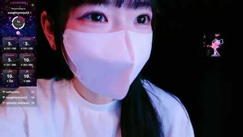 Media: A video of an East Asian woman with long black hair and fair skin, wearing a white face mask and white shirt, against a dark background, with a digital interface on the left showing a clock, weather, and a chat window.