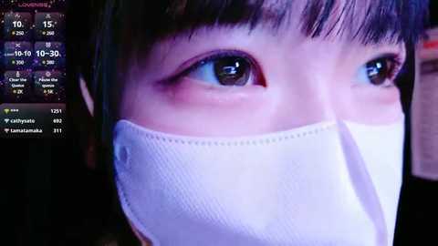 Media: A close-up video of an Asian woman with fair skin, large brown eyes, and straight black hair. She wears a white surgical mask. The background shows a dimly lit room with a smartphone displaying weather and location details.