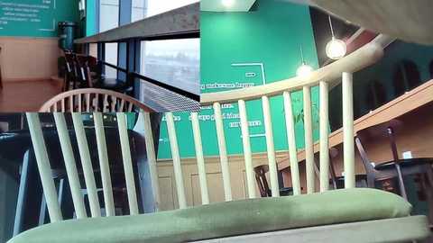 Media: Video of a modern, brightly-lit room with a green wall and wooden staircase, featuring a light beige railing and a glass window. The room includes a wooden chair and a green upholstered bench.