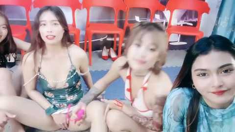 Media: Video of four Asian women with fair skin, wearing revealing lingerie, sitting on a floor with red plastic chairs in the background.