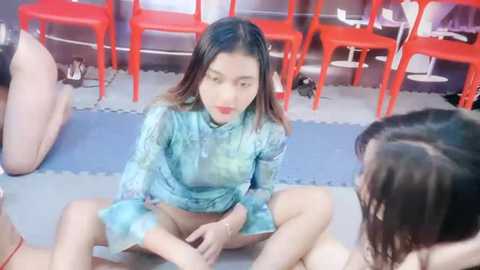 Media: A video shows a young Asian woman with long, straight hair in a blue patterned top, sitting on a mat surrounded by red stools in a modern, minimalist room.