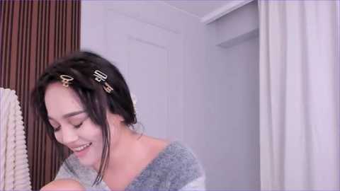 Media: Video of a young Asian woman with shoulder-length dark hair, wearing a grey off-shoulder sweater, smiling and looking down. Background features a white wall and a textured brown curtain.