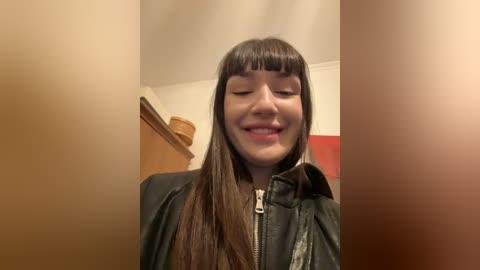 Media: Video of a young woman with long brown hair and blunt bangs, wearing a black leather jacket, smiling with eyes closed, standing indoors near wooden cabinets and a red wall.