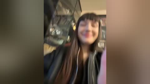 Media: A video captures a young woman with long, straight brown hair and bangs, wearing a black jacket and headphones, smiling. The background features blurred shelves with books and clothes, suggesting a cozy, possibly cluttered room.