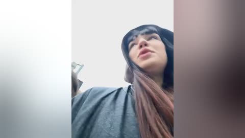 Media: Video of a young woman with long, straight, brown hair, wearing a black beanie and a grey hoodie, standing in a brightly lit room with a white wall, taken from a low-angle perspective.