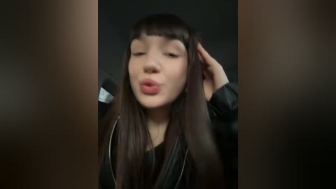 Media: Video of a young woman with long brown hair, straight bangs, and a black leather jacket, kissing her hand in a playful, exaggerated manner.