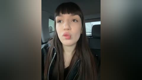 Media: Video of a young Asian woman with straight, dark brown hair and blunt bangs, wearing a black leather jacket, puckering her lips. She's seated in the backseat of a car, with blurred background showing a grey interior and a window.