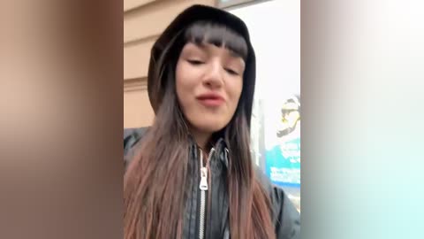 Media: Video of a young woman with straight, long, dark brown hair, wearing a black beanie and jacket, standing in front of a storefront with a blue sign. She has a neutral expression, eyes closed.